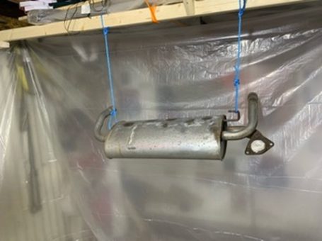 Metal exhaust muffler hanging from strings in a workshop setting.