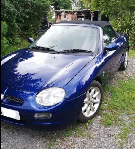 Blue convertible sports car parked on a gravel driveway surrounded by greenery.