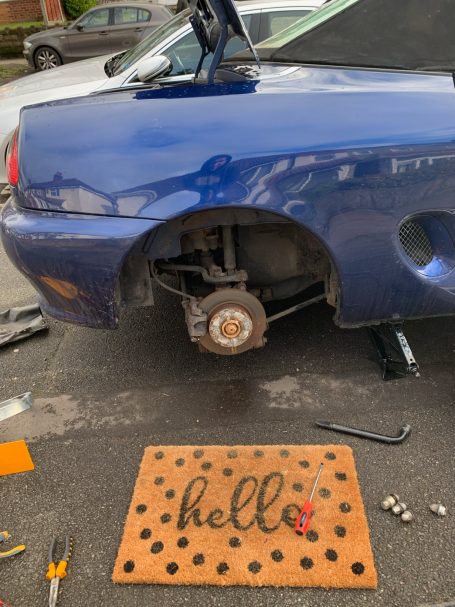 Blue car with a rear wheel removed, parked on a drive with a "hello" doormat nearby.