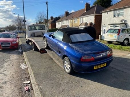 Blue convertible car being loaded onto a tow truck on a residential street.