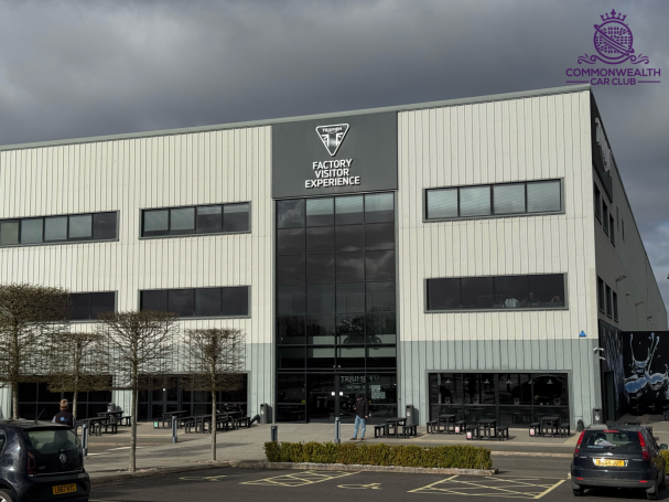 Modern building with large windows and logo, surrounded by trees and parked cars.