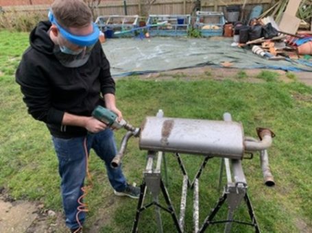 Man wearing a visor uses a drill on a muffler placed on a workbench outdoors.