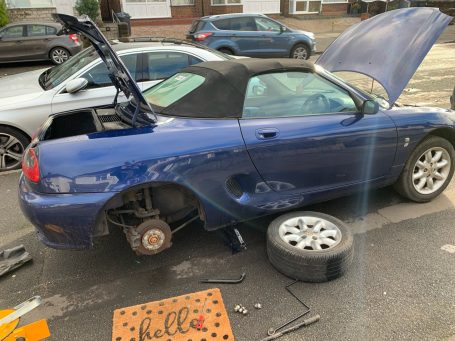 Blue convertible car with its hood and boot open, missing front wheels, parked on a street.