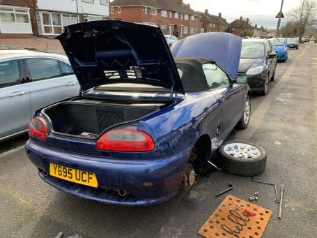 Blue convertible car with the hood open, parked on a street, missing rear wheels.