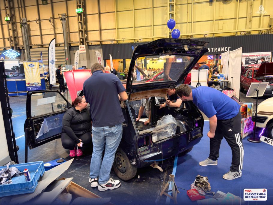 Three people work on dismantling a car in an exhibition space with tools nearby.