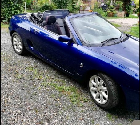 Blue convertible car parked on gravel, with the roof down and lush greenery in the background.