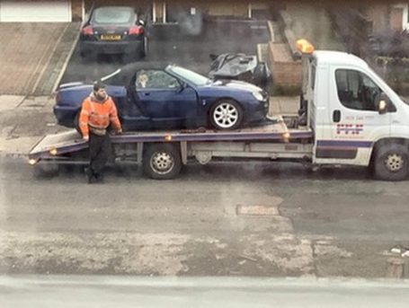 A tow truck loading a blue car with a person in an orange jacket seated nearby.