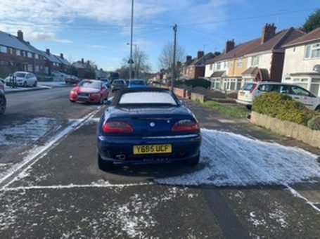 A blue car parked on a snowy street, with houses and other vehicles in the background.