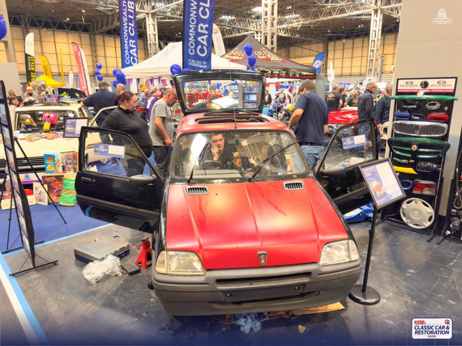 Red car displayed with doors open at an exhibition, surrounded by attendees and vendor booths.