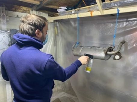 A person wearing a mask works on a metal exhaust part in a sheltered area.